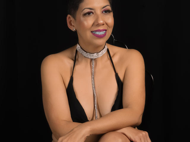 JuliaNewell with short black hair, purple lipstick, and black halter top, showing small tits, wearing a diamond choker, arms crossed, smiling, against a black background.