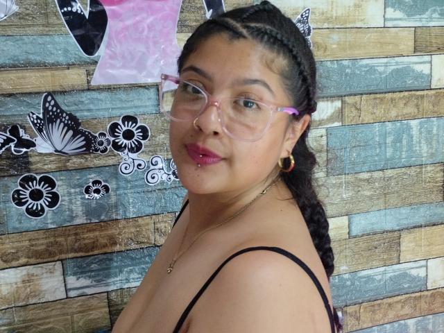 BrunetteLiaSex with glasses, pink frames, braided hair, gold earrings, pink lipstick, and black strap top, showing her tits, smiling, against a wooden wall with butterfly and flower decals.