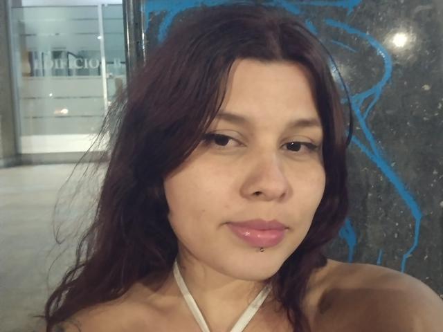 SparklesRomance hat braunes Haar, rosafarbene, glänzende Lippen, ein Lippenpiercing und trägt ein weißes Top mit Halter, mit einem leichten Lächeln, auf einem Selfie..