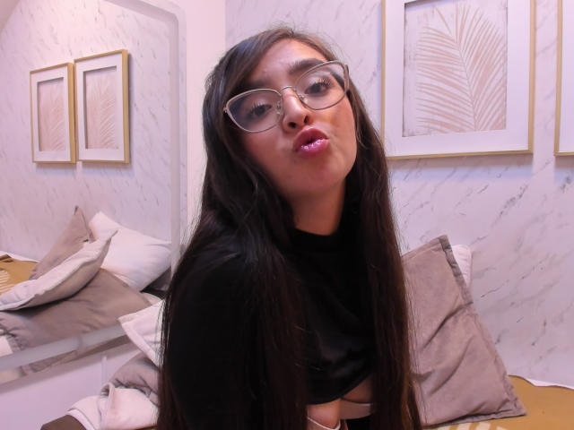 Eva McAllister mit langem, schwarzem Haar, Brille und pinkem Lippenstift, leicht schmollend, trägt ein schwarzes Oberteil, das einen kleinen Teil ihrer Brust zeigt, in einem gemütlichen Raum..
