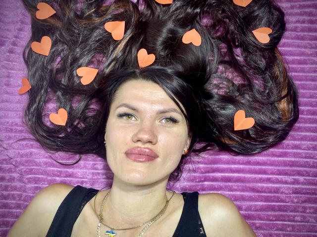 MilaLoona's face with puffy pink lips, dark brown wavy hair spread out, orange heart stickers, wearing a black top, gold necklace, and a blue pendant, against a purple textured background.