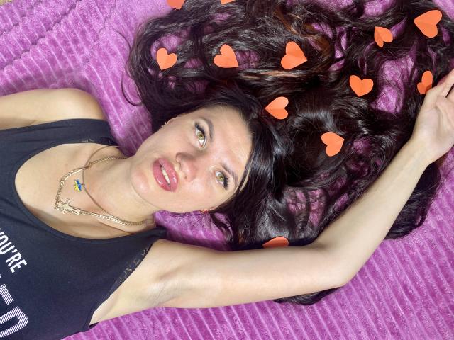 MilaLoona with long black hair, wearing a black tank top, lying on a purple surface, arms above head, orange heart emojis on her hair, slightly parted red lips.