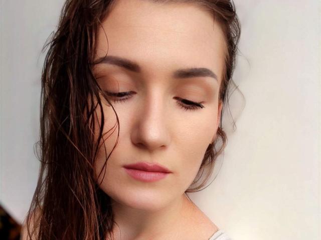 Anny's face with wet brown hair, closed eyes, and subtle pink lipstick. Her eyelashes are long and she has a calm, slightly downcast expression. Soft, fair skin.