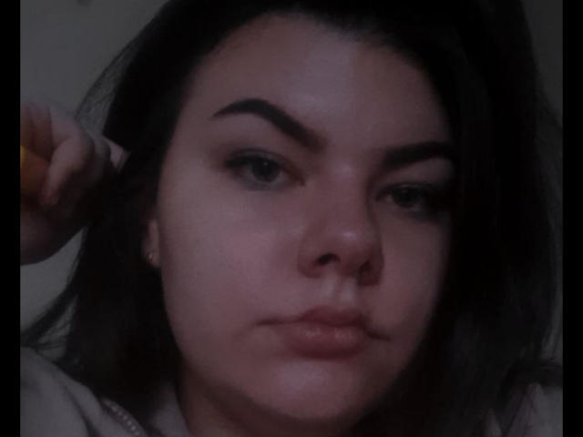O rosto de TinaMoore, com sobrancelhas escuras, lábios ligeiramente entreabertos e cabelo escuro em uma selfie com pouca luz. Sua mão direita segura um objeto amarelo perto de sua orelha..