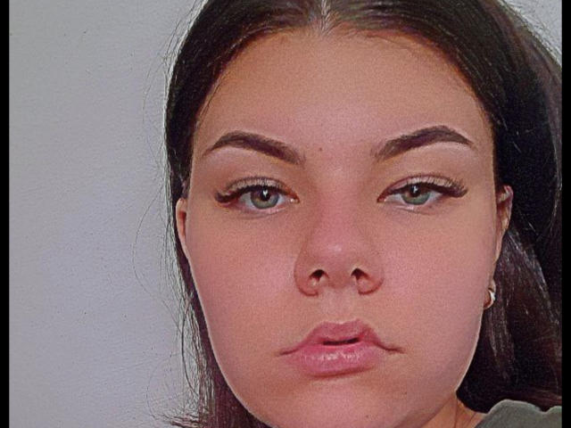 Tinas Moores Gesicht zeigt volle, rosafarbene Lippen mit einem leichten Schmollmund, grüne Augen mit langen Wimpern und dunkelbraunes, glattes Haar. Sie hat einen hellen Hautton und dicke Augenbrauen..