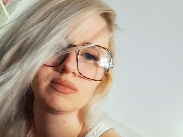 ValeOana, con i capelli biondi, gli occhiali a tartaruga e le labbra rosa, che mostra un leggero sbuffo. Ha una piccola lentiggine sul naso. Indossa una maglietta bianca..
