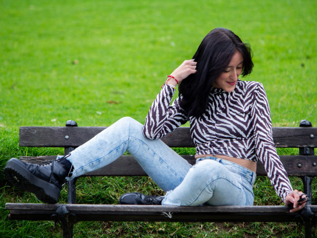 Envyv sitzt auf einer hölzernen Bank, die Beine gespreizt, und trägt ein zebra-gestreiftes Oberteil, helle blaue Jeans und schwarze Stiefel. Sie berührt ihr Haar und lächelt..