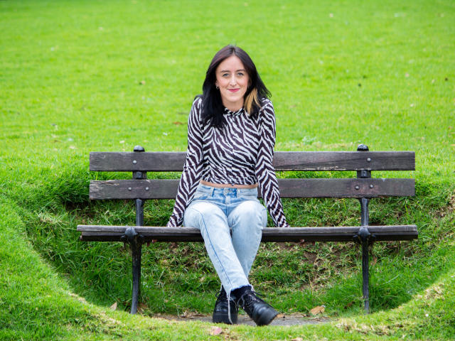 Uma mulher com cabelo preto, vestindo uma blusa com estampa de zebra, jeans azuis e botas pretas, sentada em um banco de madeira, sorrindo, com as pernas cruzadas, e um fundo com grama..