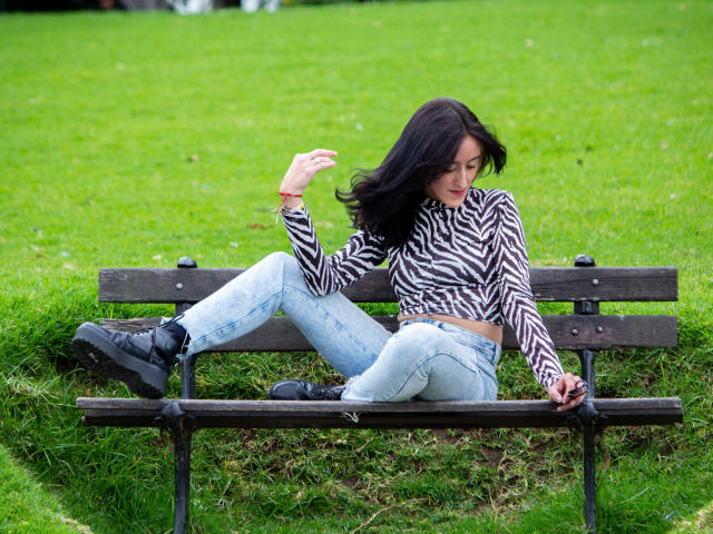 Envyv with black hair, wearing a zebra-striped top and light blue jeans, sits on a dark wooden bench, legs crossed, right arm raised, looking down, in a grassy park.
