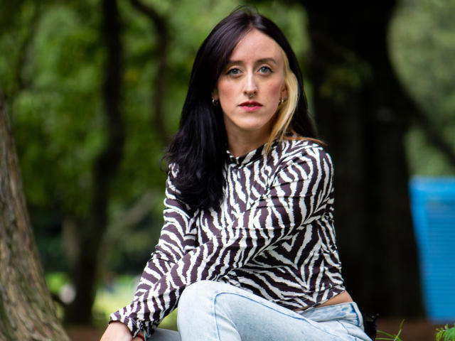 Envyv tiene el cabello negro y rubio, lleva una camisa con estampado de cebra y jeans azul claro, está sentada contra un árbol, mirando ligeramente hacia un lado con una expresión neutra..