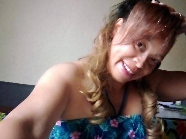 Rahel69, con la pelle scura, sorridente, indossa un top blu a fiori senza spalline, ha i capelli biondi ricci e delle mollette nere per i capelli, ha un braccio alzato, mostrando il seno e un viso carino..