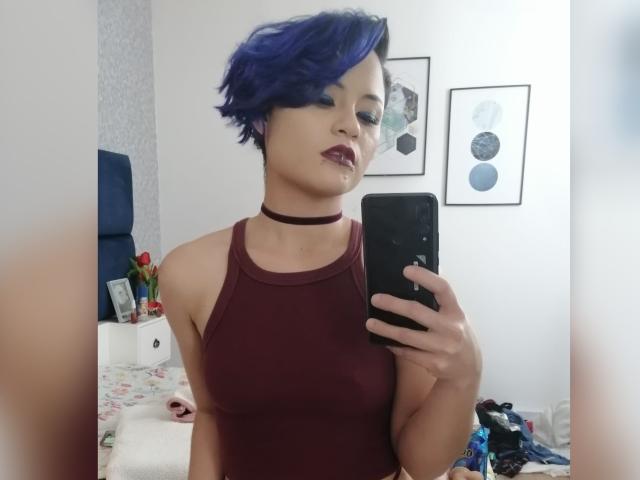 CassieJones with blue pixie hair, dark lipstick, and a maroon choker, takes a mirror selfie in a bedroom, wearing a maroon tank top, showing her small tits.
