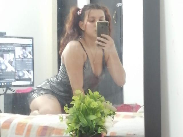 SparklesRomance mit lockigem, braunem Haar in zwei Pferdeschwänzen, trägt ein glänzendes, graues Kleid, kniet auf einem Bett und macht ein Selfie im Spiegel, wobei ihr Hintern und ihre kleinen Brüste zu sehen sind..