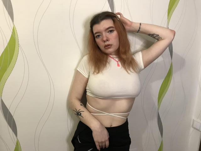 Amelia Aley, con el cabello teñido de rosa, un top corto blanco que deja ver los pezones, pantalones negros, un tatuaje en el brazo izquierdo, con la mano en el cabello, de pie frente a una pared con un patrón ondulado de color verde y gris..