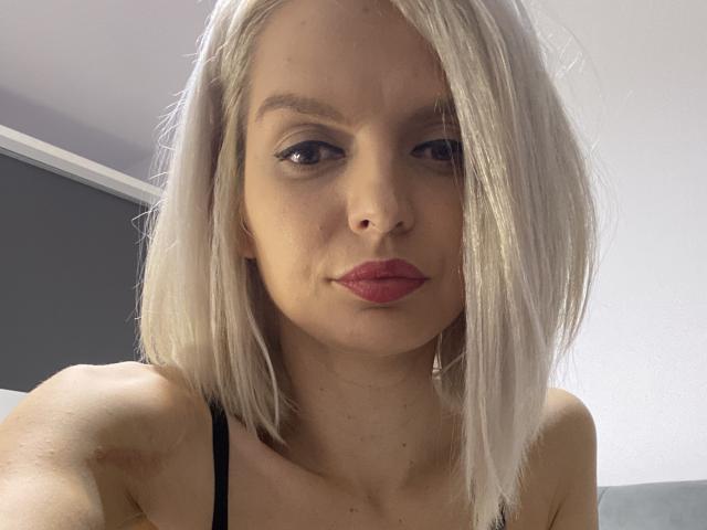 DouceAmelia has pale skin, straight platinum blonde hair, black eyeliner, and red lipstick. She's wearing a black strap, and her boobs are partially visible. She's staring at the camera.