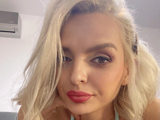 DouceAmelia has blonde hair, blue eyes, red lipstick, and a nose piercing. She's looking at the camera with a slight smile. Her tits are not visible.