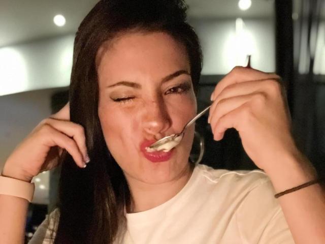 Liliysweet has dark hair, winks with one eye, and eats yogurt with a spoon. Her red lips are slightly smeared with yogurt. She wears a white shirt.