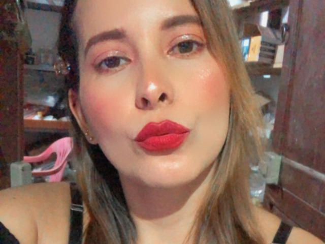 SaritaVelez with puffy red lips, glossy skin, and brown hair, pouting her full, glossy pink lips, wearing a black top, looking at the camera with pink eyeshadow.