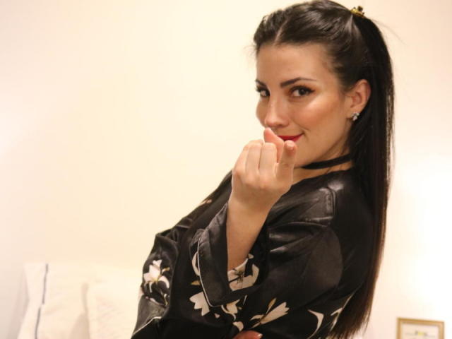 Liliysweet with dark hair in a ponytail, wearing a black floral top, biting her red-lipped finger, looking over her shoulder with a playful smile.