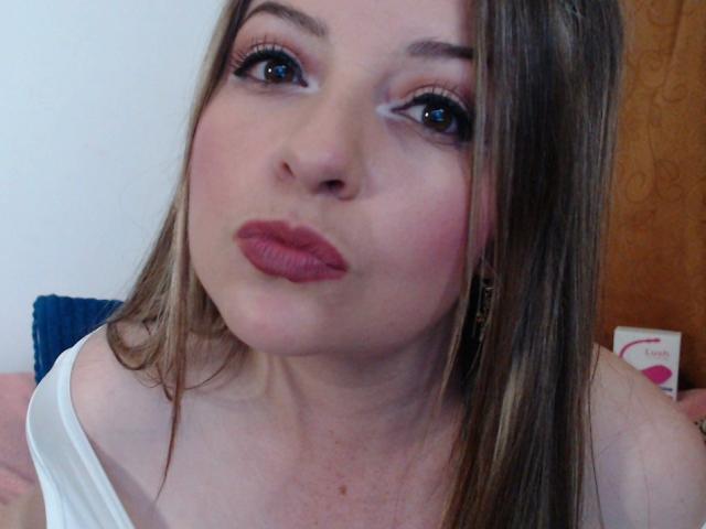 NatalyHami with pouty pink lips, brown eyes, and long brown hair, wearing a white off-shoulder top, making a cute, playful face. Boobs partially visible.