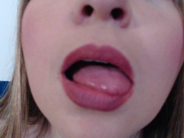 NatalyHami's face, showing her pink tongue slightly out, with full red lips and fair skin, blurred nose, and blonde hair on the sides. Her mouth is slightly open.