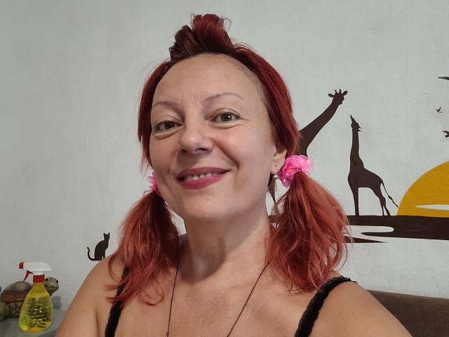 BarbaraBowie con due codini rossi, elastici rosa per capelli, che sorride, indossa una canottiera nera, ha un seno piccolo, pelle chiara, rossetto rosso, una parete bianca, una bottiglia spray gialla e un'opera d'arte a tema giraffa sulla parete..