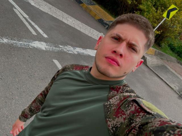 SebastiianMaxime, con cabello castaño corto, una camisa verde y una chaqueta de camuflaje, mostrando una expresión de dolor, está de pie en una carretera con líneas blancas y una señal de tráfico amarilla..