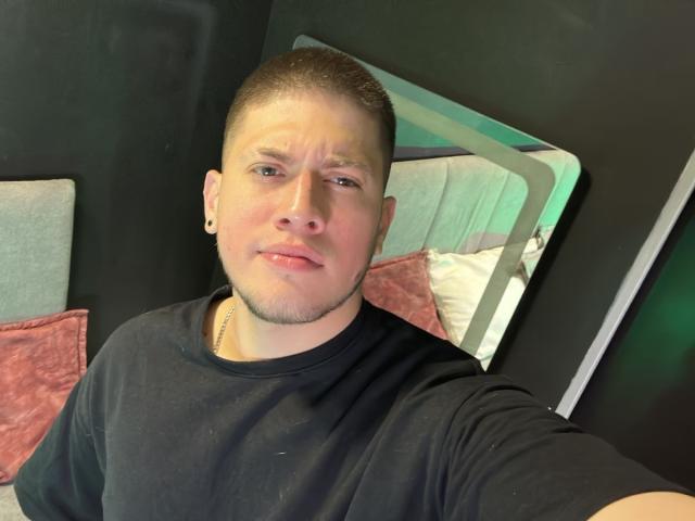 SebastiianMaxime tiene un corte de pelo muy corto, una camisa negra, un pequeño aro en la nariz, una leve sonrisa y perforaciones visibles en las orejas. Su rostro muestra una expresión sutil y relajada..