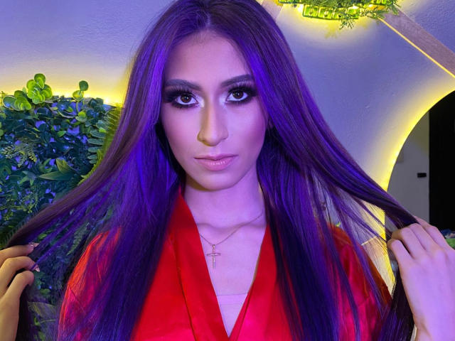 JuliaJobs with purple hair, red robe, holding her hair, wearing heavy makeup, cross necklace, looking at camera, purple and yellow lighting, green plant in background.