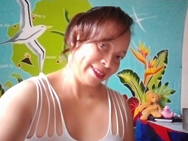 Rahel69 with brown hair, smiling, wearing a white, cut-out, tank top, showing her small, perky tits, in a brightly colored room with a stuffed toy and bird painting.