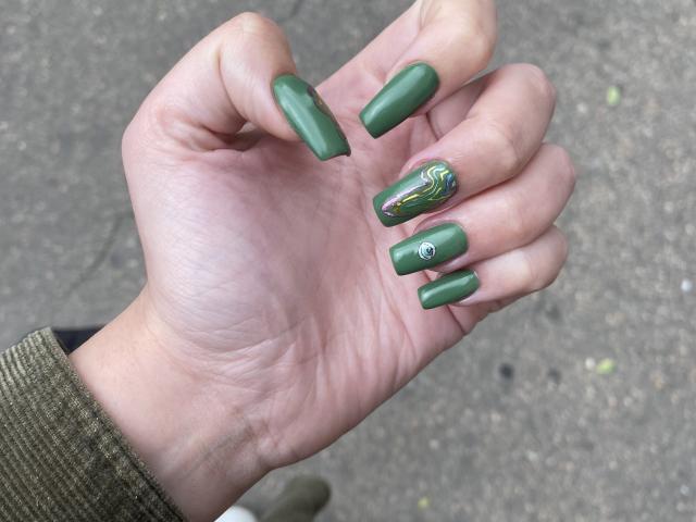 La main de Rubbi, avec des ongles verts en forme d'amande, dont un orné d'un motif coloré et tourbillonnant ainsi que d'une petite gemme, sur un fond gris texturé. Elle porte une manche verte et texturée..