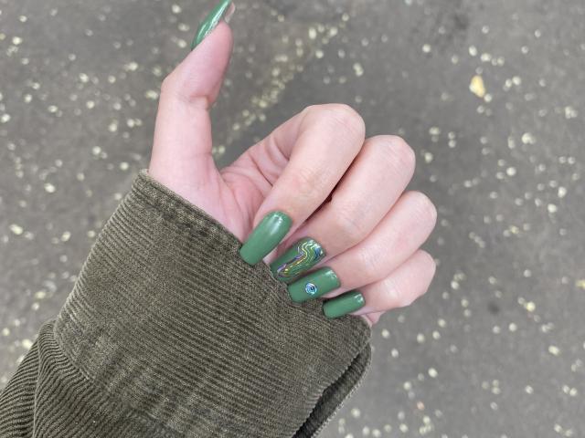 Rubbi's hand with green, long, almond-shaped nails, featuring a small blue gem and green design on the middle nail, wearing a green corduroy sleeve.