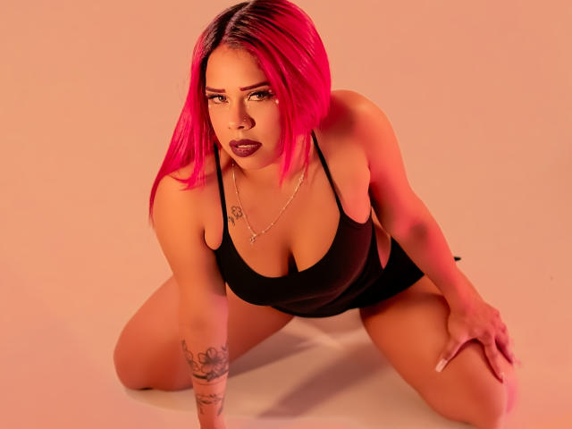 Valentina, com cabelo rosa, top preto e tatuagens, ajoelhada no chão, com as mãos nos joelhos, olhando para cima com batom escuro, mostrando seios grandes e decote, em uma luz laranja suave..