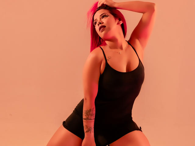 Valentina, con cabello rosa, camiseta negra y pantalones cortos, arrodillada, con una mano en el cabello, tatuaje en el brazo, luz roja, busto grande, sexy, pose segura..