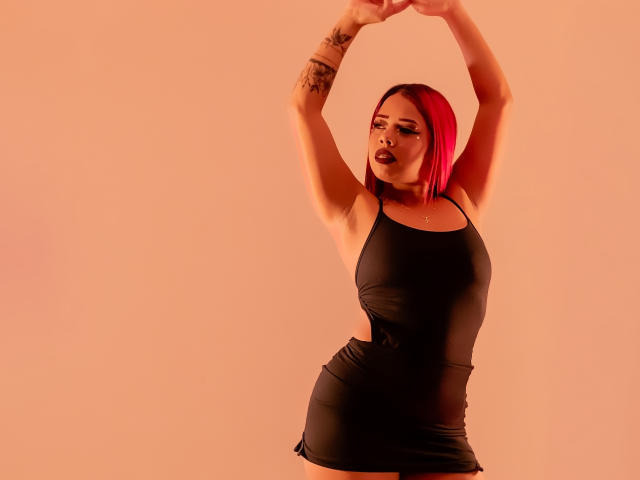 Valentina, avec une coupe de cheveux courte rose, un débardeur noir, les bras levés, un tatouage sur l'avant-bras droit, portant du rouge à lèvres foncé, debout, sur un fond de couleur pêche, montrant ses seins et une petite jupe noire..