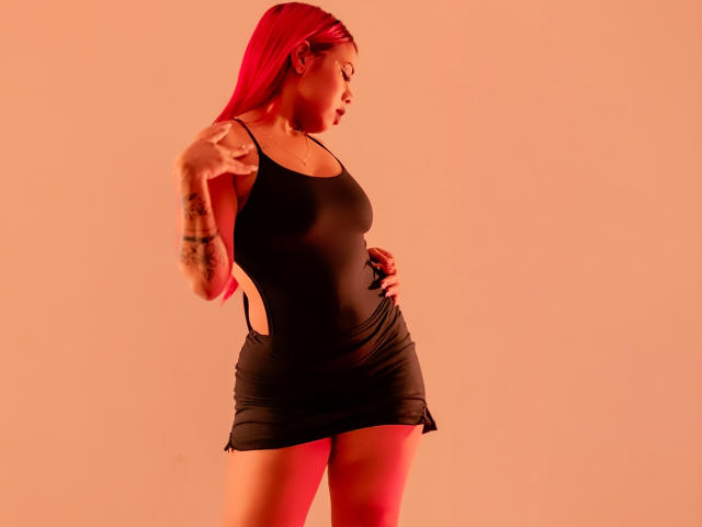Valentina, com cabelo ruivo, vestindo um vestido preto justo, exibindo suas nádegas grandes e pequenos recortes laterais, apontando com o braço tatuado, em pé contra uma luz rosa-avermelhada..