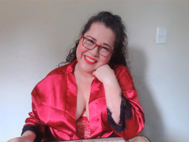 Loretta Sanchez, com cabelo preto encaracolado, óculos vermelhos e um robe de cetim vermelho, sorrindo, mostrando o colo, lábios vermelhos e a mão no rosto, sentada. Seios visíveis, com um sutiã vermelho de renda por baixo..