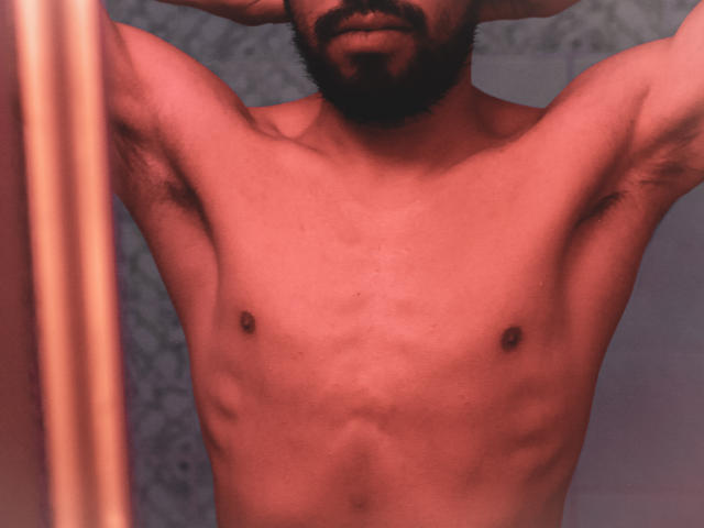 AngelsScott, con il suo torace muscoloso e scoperto, piccoli capezzoli visibili, peli sotto le ascelle e barba scura, braccia alzate, testa parzialmente fuori dall'inquadratura, in una foto sfocata con tonalità rosse..