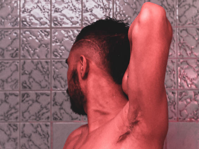 AngelsScott, com um lado da cabeça raspado, barba e cabelo molhado, levantando o braço para mostrar os pelos da axila, em um banheiro com iluminação vermelha..