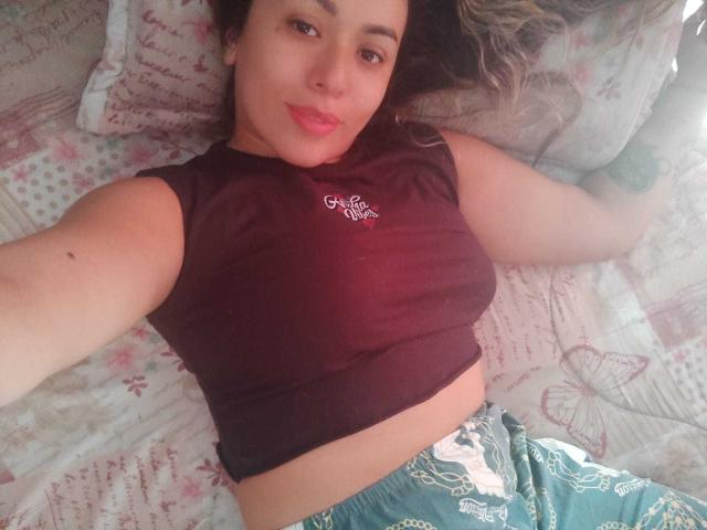 Joanallen a des seins de taille moyenne, porte une chemise marron, un short vert, est allongée sur un lit, sourit, a les cheveux bruns, du rouge à lèvres rose, une peau claire, un tatouage sur le bras, c'est un selfie décontracté, elle a les yeux marron clair, elle a l'air détendue..