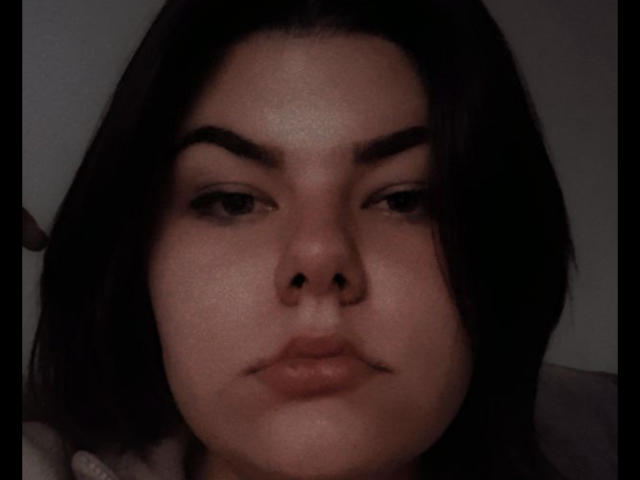 TinaMoore has dark, wavy hair, thick eyebrows, and a small mustache above her slightly parted, glossy lips. Her skin is fair with a subtle sheen.