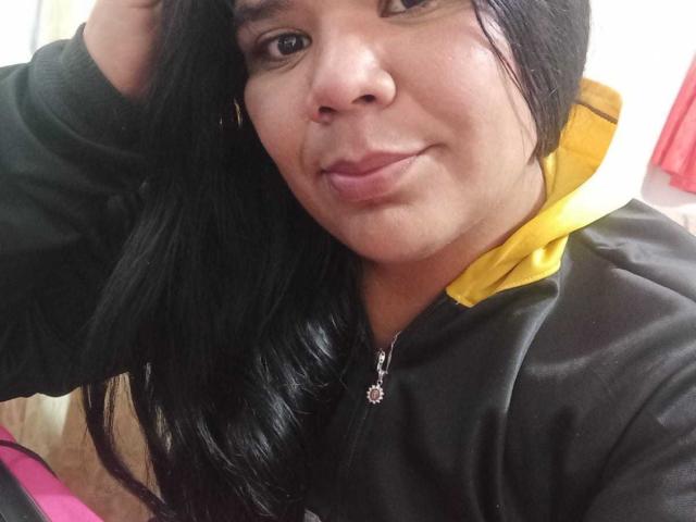 LiAngel a une peau marron moyen, des cheveux noirs, et elle porte un sweat à capuche noir avec une fermeture éclair et un col jaune. Elle regarde l'appareil photo avec un léger sourire. Elle porte un collier..