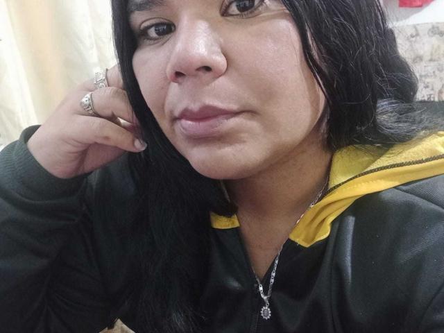 LiAngel con labios carnosos, cabello negro, chaqueta verde, capucha amarilla, anillo en la mano izquierda, collar y una ligera sonrisa. Su rostro está centrado, y una mano le toca el cabello..