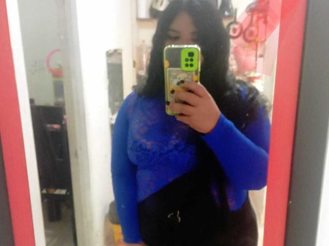 LiAngel with dark wavy hair, wearing a blue lace top, black pants, holding a green phone in front of a mirror, reflecting her upper body and face.