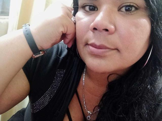 LiAngel tiene una piel marrón media, cabello negro y rizado, y lleva una camisa negra con detalles brillantes, un collar de plata y una pulsera negra. Está apoyando su barbilla en su mano..