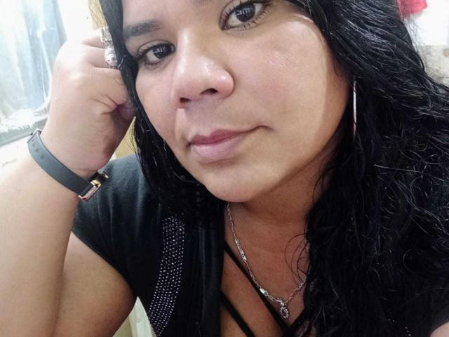 LiAngel has dark wavy hair, brown eyes, and a slight smile. She wears a black shirt, silver necklace, and hoop earrings with a black wristband. Her pussy is not visible.