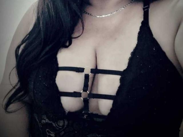 Los senos de LiAngel en un sujetador negro de encaje con un arnés de tiras, mostrando el escote. Cabello negro largo, collar de plata, piel clara, iluminación suave, ligera sombra..