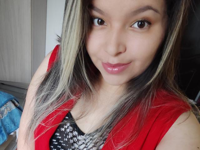 Myalewwis heeft een lichtbruine huid, glanzende roze lippen en zwart haar met blonde highlights, draagt een rode top over een zwart-wit gestreept shirt en glimlacht lichtjes..