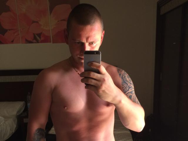 MichaelHero, sin camisa y con la cabeza afeitada, sosteniendo un teléfono, tomando una selfie en el espejo. Tiene un tatuaje en su brazo derecho, pezones visibles y una expresión seria..