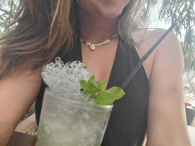 WildBlondy69 with ice in a tall glass, green mint, straw, black halter dress, shell necklace, brown wavy hair, visible chest, and a relaxed outdoor setting.